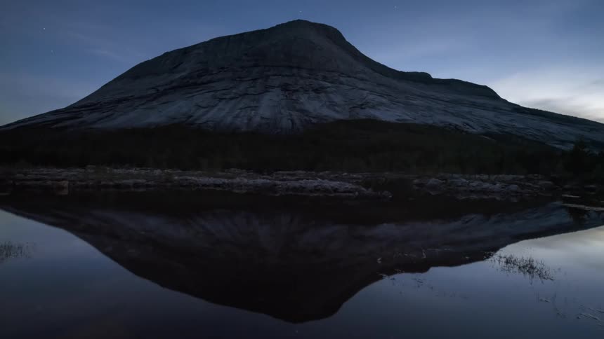 Rocky mountain on the edge of a lake that reflects the dawn and dusk sky along with an aurora borealis