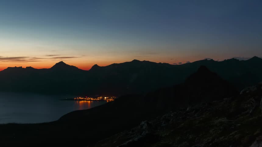 Twilight landscape with silhouettes of mountains against a gradient blue sky with aurora borealis and city lights visible in the distance on the edge of a body of water