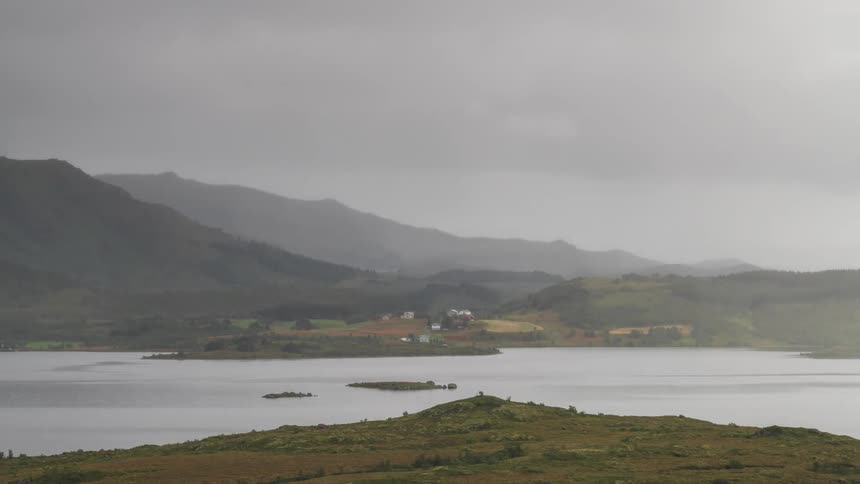 Fog rolls over a lake and a house near the shore in front of mountains