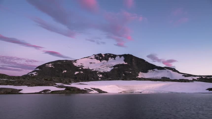 Snow covers part of a mountain behind a lake as the sun sets