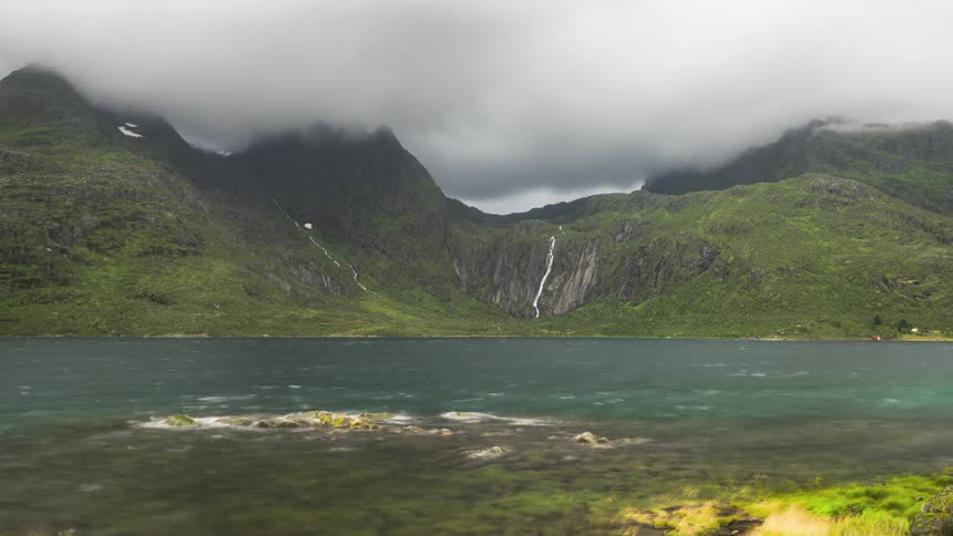 Water moves past coastal mountains partially covered by dense clouds