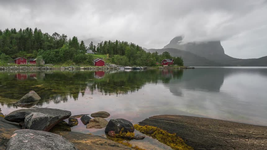 Calm lake surrounded by mountains and small houses on a dense overcast day