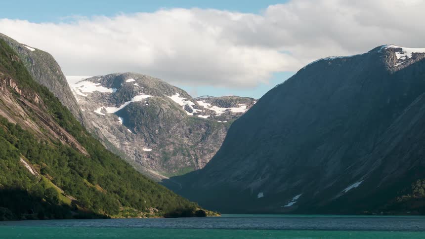Scenery of a sea canal with turquoise blue water surrounded by mountains with snowcapped peaks and clouds passing by