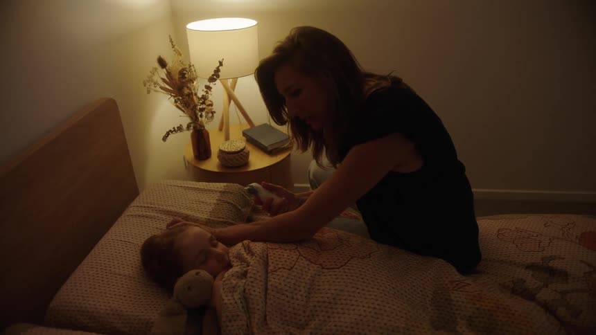 Mother sits on bed while taking the temperature of her daughter