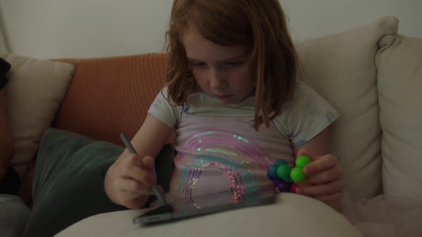 Little girl sitting on the couch playing on her ipad as her mom sits nearby