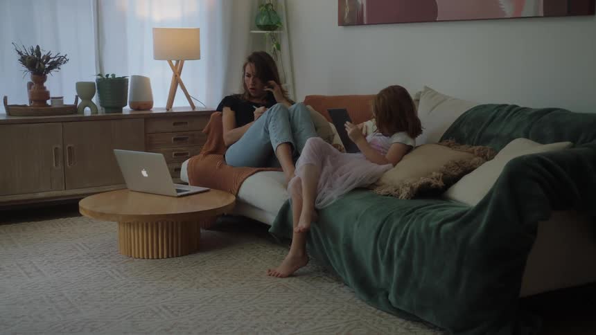 Woman working on a laptop and talking on the phone while sitting on the couch next to her daughter who is using a tablet