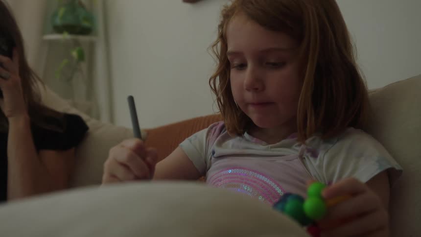 A mother speaks on a smartphone while her daughter sits next to her on a couch drawing