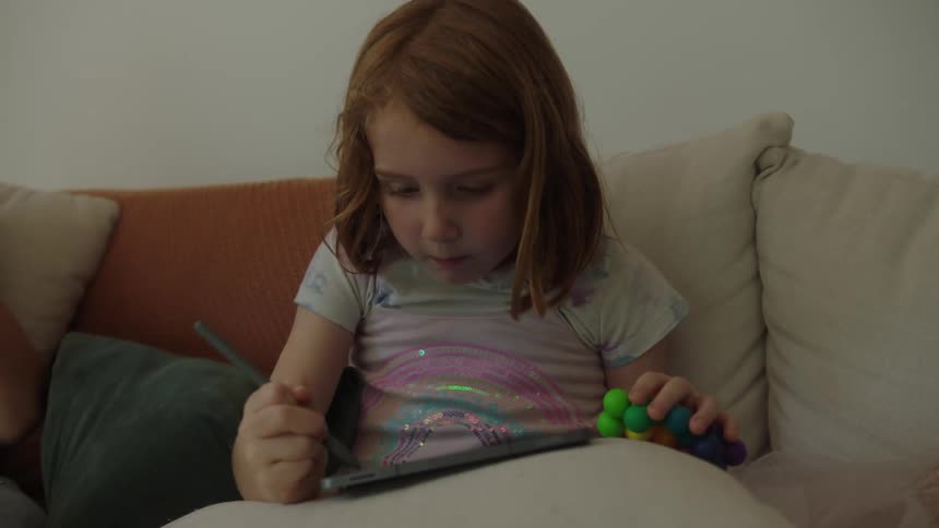 Young girl sitting on the couch playing on her ipad as her mom sits nearby