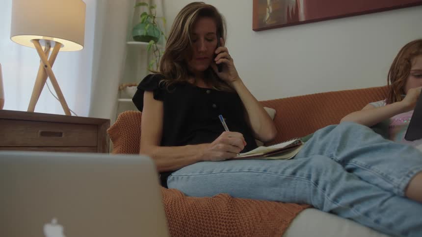 Mother sits next to her daughter on a couch while taking notes and talking on the phone