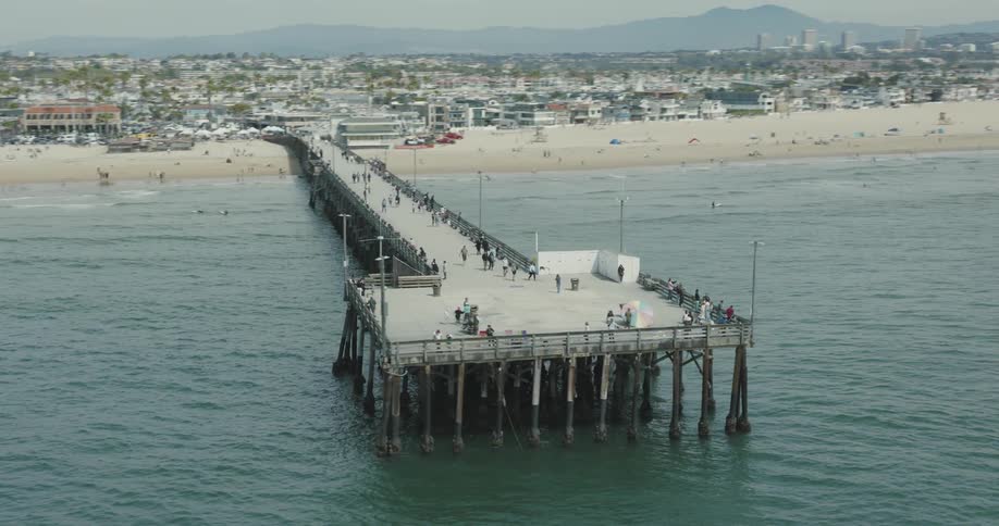 Aerial panorama of an extensive dock that reaches the sea from the beach with hundreds of people and a city in the background
