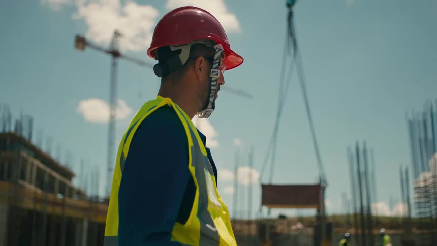 A construction worker in a yellow hard hat and reflective vest is controling a crane on a construction site