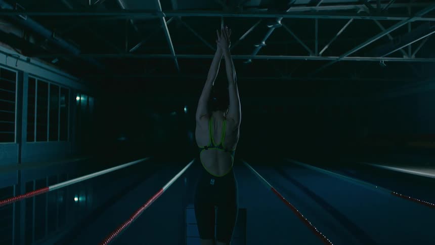 Woman with her back stretching her arms upwards and climbing a ladder in front of an indoor pool at night
