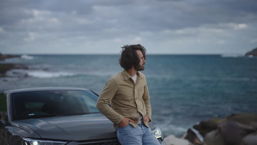 Man leans against a car parked on shore and looks out toward the water