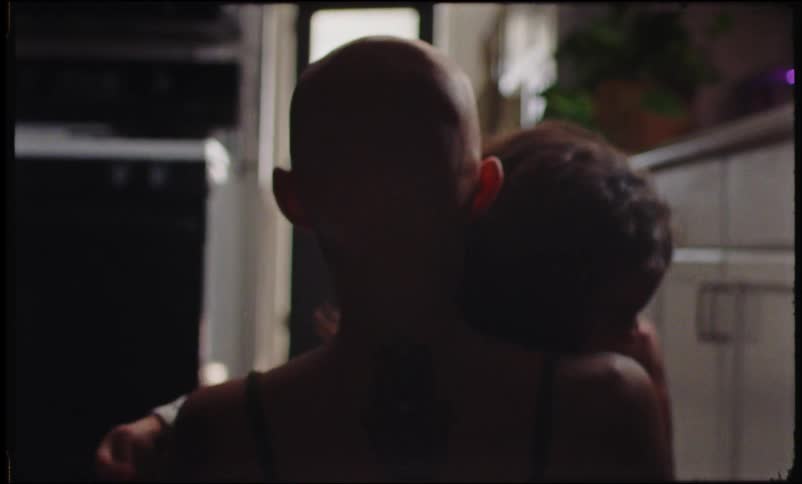 A bald woman is sitting on the floor with her back turned and then a child hugs her from the front