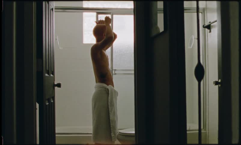 A tattooed bald woman with scars on her chest shaves her head in the bathroom