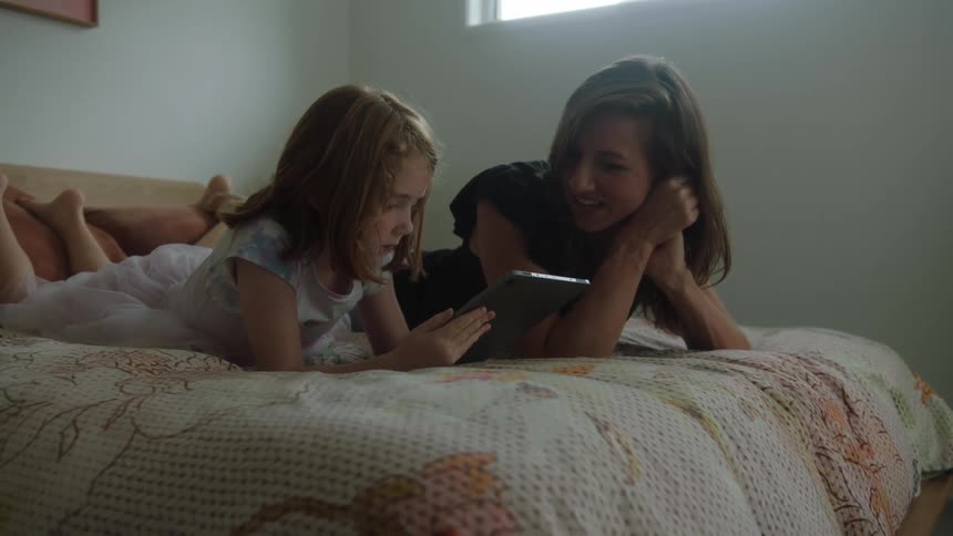 Mother lying on bed with daughter smiles at the child as the girl uses a tablet