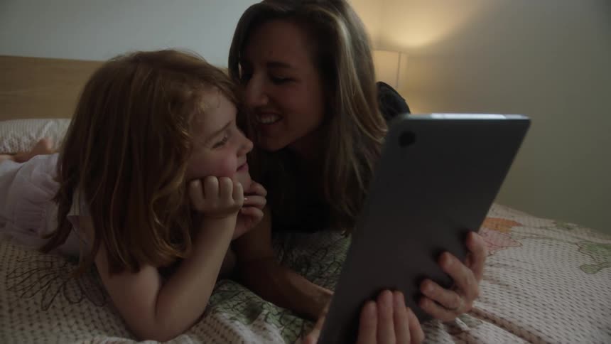A mother and daughter lying on a bed reading on a tablet