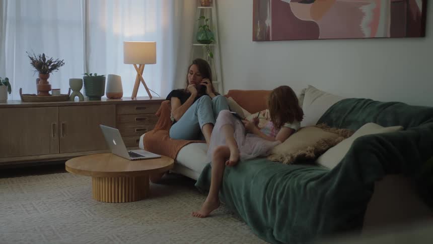 Cozy living room with mother talking on the phone and daughter using a tablet while relaxing on the sofa