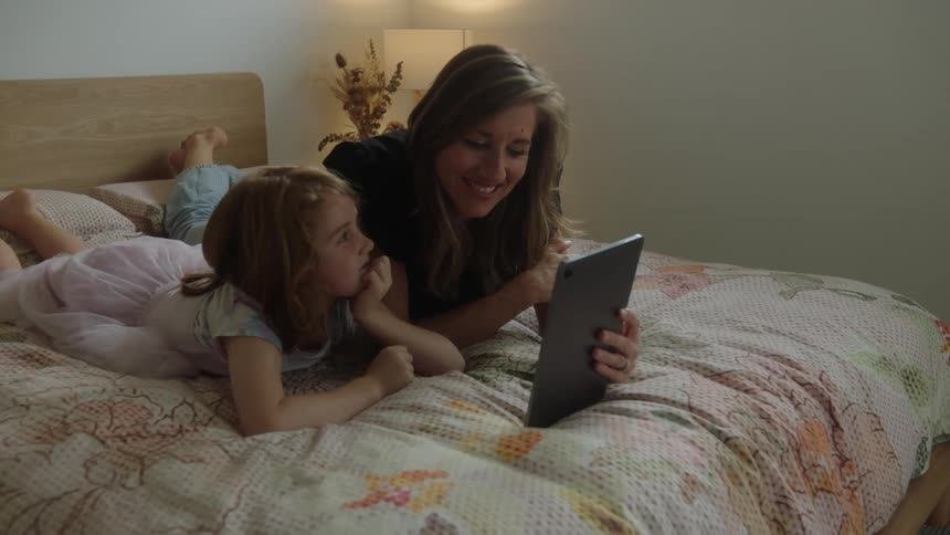 Mother sharing a tablet while lying on the bed with her daughter leans over to kiss the child