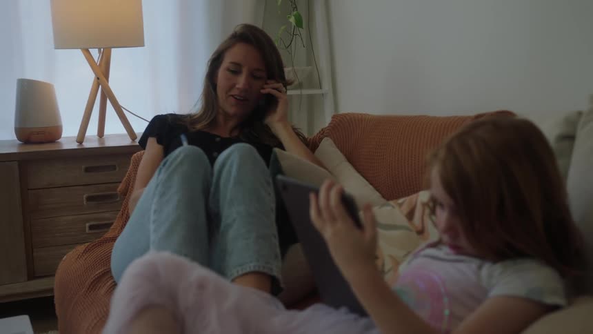 Mother and daughter relaxing on the couch at home using phones and tablets