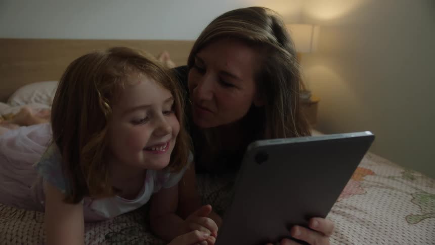 A mother and daughter look at a tablet and laugh and kiss in a bedroom