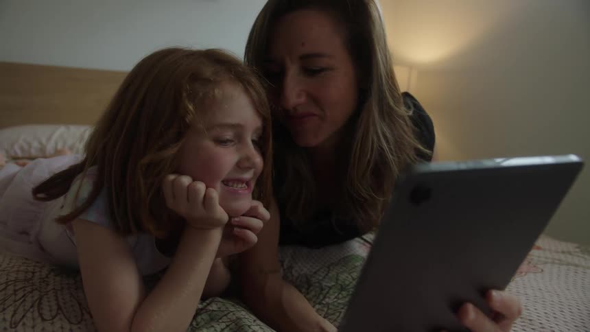 Mother and daughter lying on a bed looking at a tablet