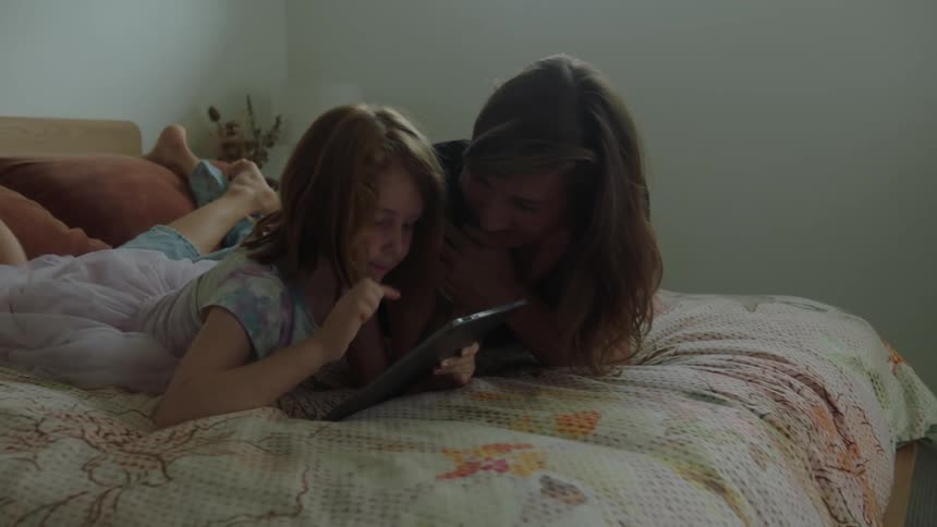 A daughter uses a tablet while lying in bed with her mother