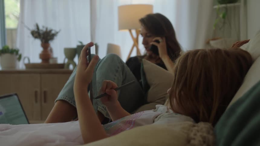 Little girl drawing on a tablet and her mother next to her talking on the phone