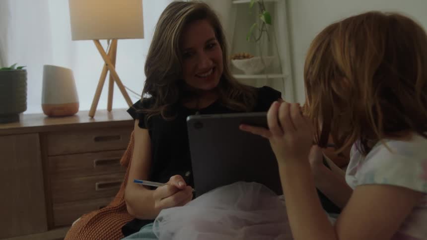Little girl sitting on the sofa drawing on a tablet and showing it to her mother who is next to her