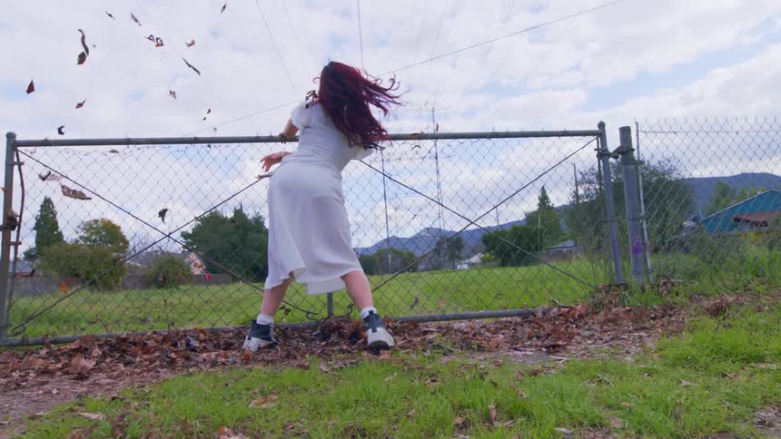 Woman in an openair environment next to a fence dancing and throwing leaves into the air