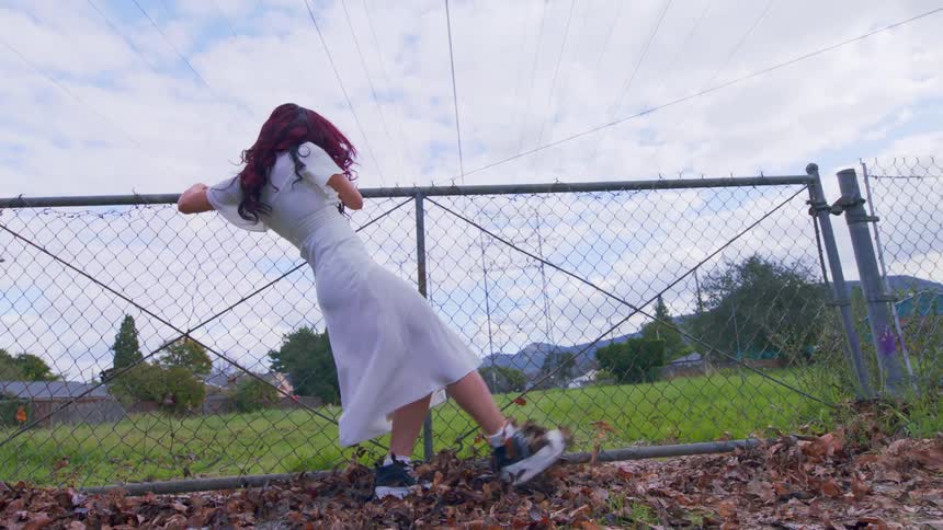 Woman hanging onto a gate leading to a fenced field kicks her foot high