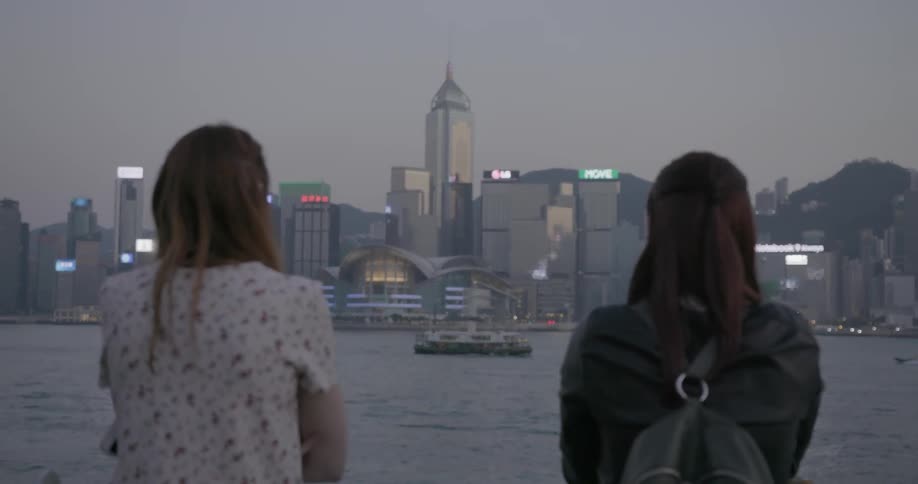 Two people sit before a city across the water one is taking pictures