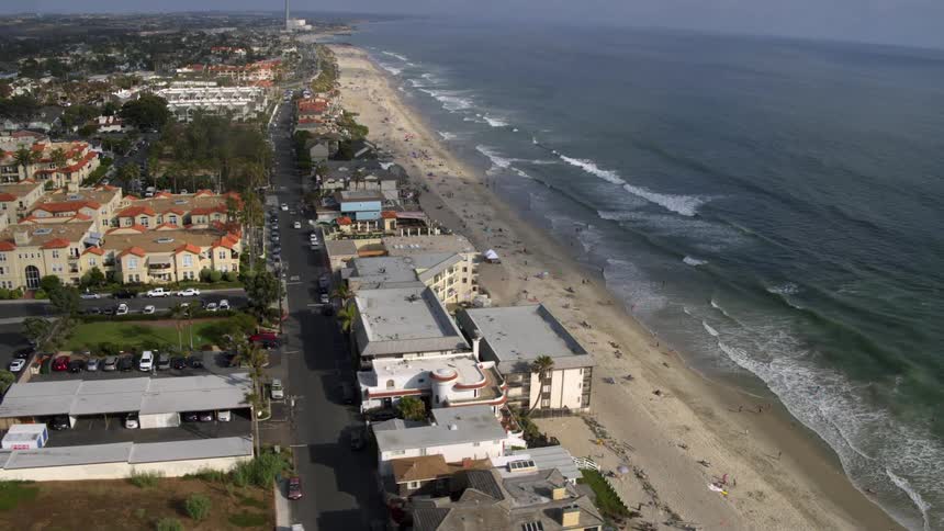Aerial view of a long beach with gentle waves crashing on the shore and a row of buildings along the shore