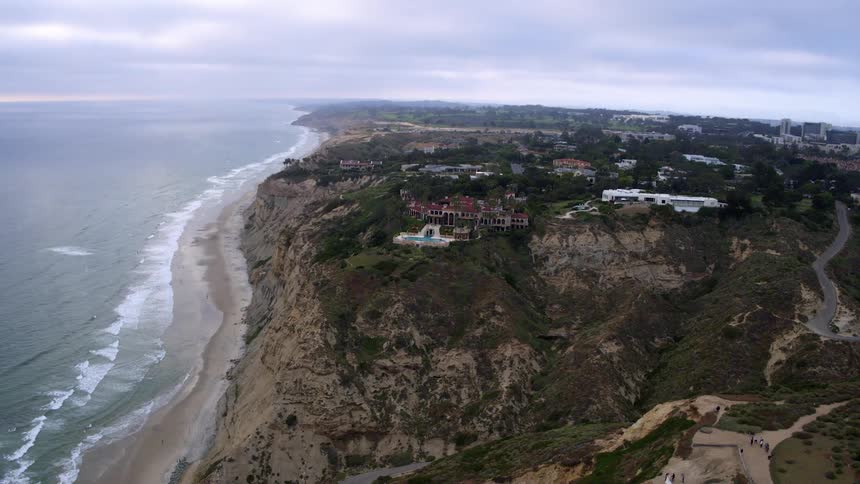 Aerial view of a coastal cliff with vegetation next to a long beach and some luxury buildings at the top