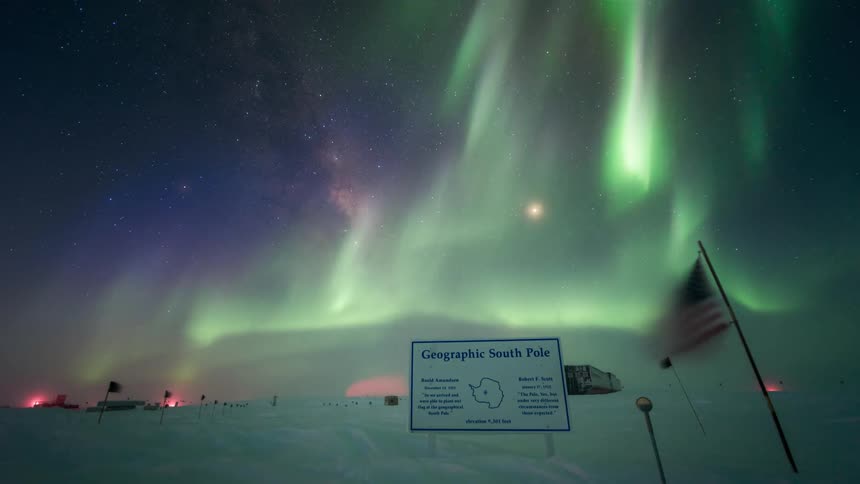 Southern aurora over the geographic south pole with a starry sky and shades of green and purple illuminating the night sky and a sign indicating the location