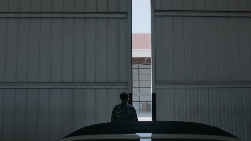 A man stands in front of two large doors which slowly open to reveal another building across the way