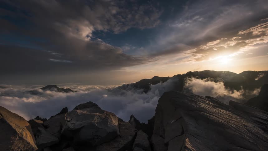 Sea of dense clouds passing above mountain peaks with the sun on the horizon