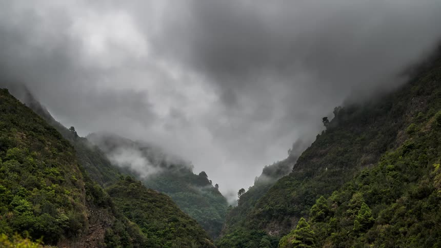 A mountainous region with a valley between them and many trees in the mountains and there are white and gray clouds flying over the place