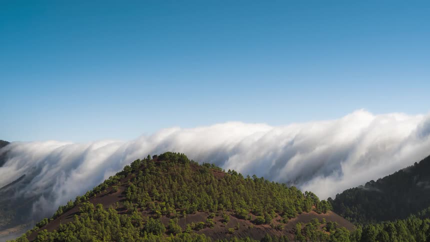 Mountainous landscape with green trees and low clouds passing against a blue sky