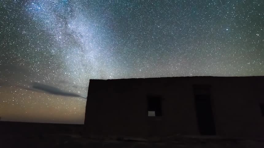 A night sky filled with rapidly moving clouds stars and the milky way galaxy with a solitary building silhouetted in the foreground