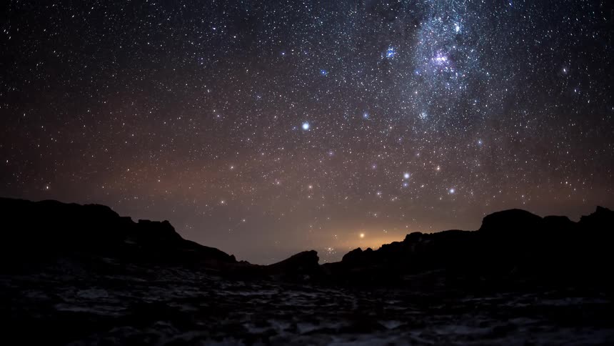 Starry night sky and part of the milky way visible above a dark silhouette of uneven terrain
