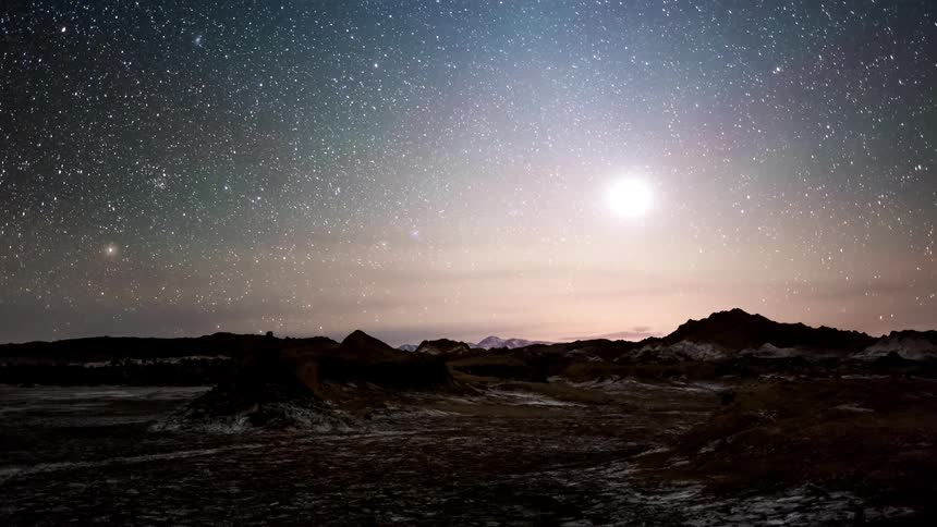 Night landscape with a starry sky and the bright moon above a rocky terrain with the sky turning shades of purple and the sun rising over the horizon