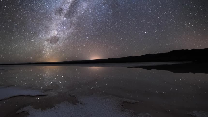 Lake with ice surrounded by silhouette of mountains that reflects the sky with the milky way