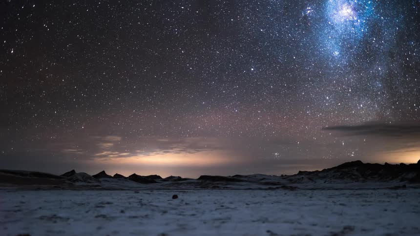 Starry night sky over an arid terrain with mountains in the background and the ground covered in snow