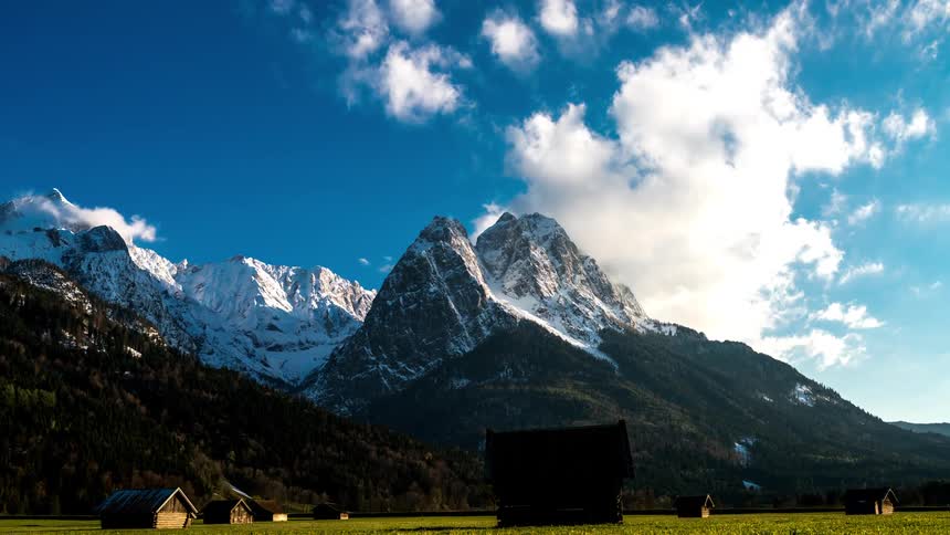 A green field with small structures against snowcapped mountains clouds rapidly moving in the sky