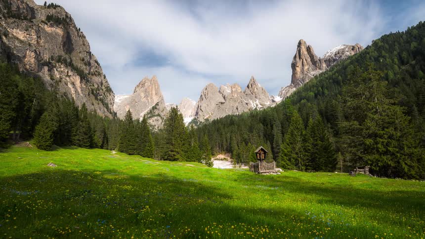 Green field with wildflowers a small wooden chapel and imposing mountains in the background under a partly cloudy sky