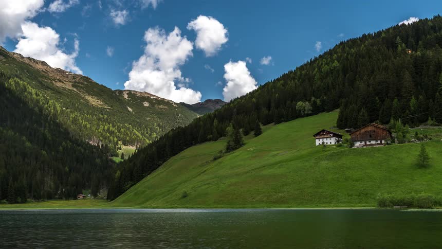 Natural scenery with a lake in the foreground surrounded by green hills and a few scattered houses under a blue sky with clouds