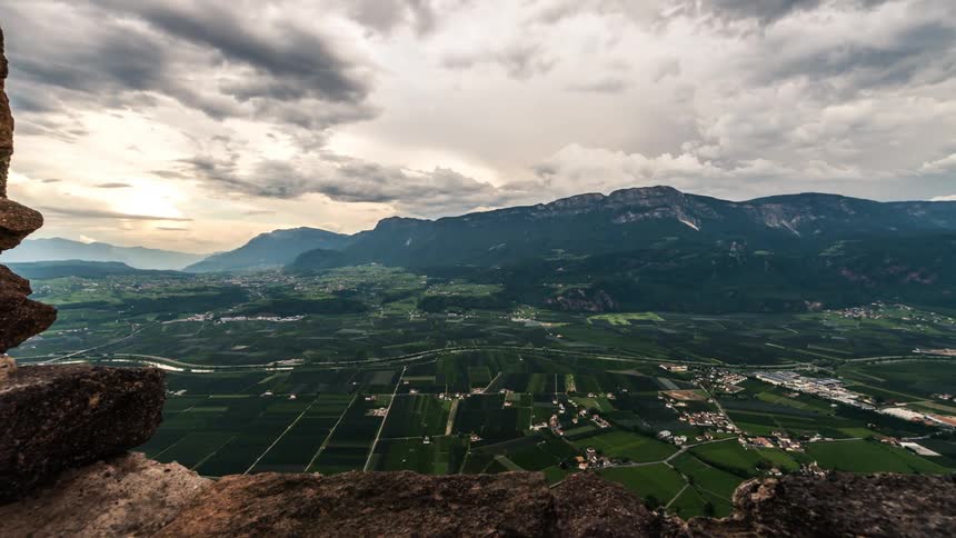 Scenery of rocks in the foreground overlooking a panorama of a village and greenish mountains against a cloudy sky