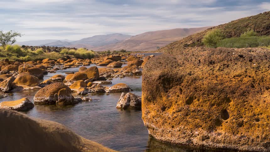 River flowing between rocks in a natural environment surrounded by mountains against a partly cloudy blue sky