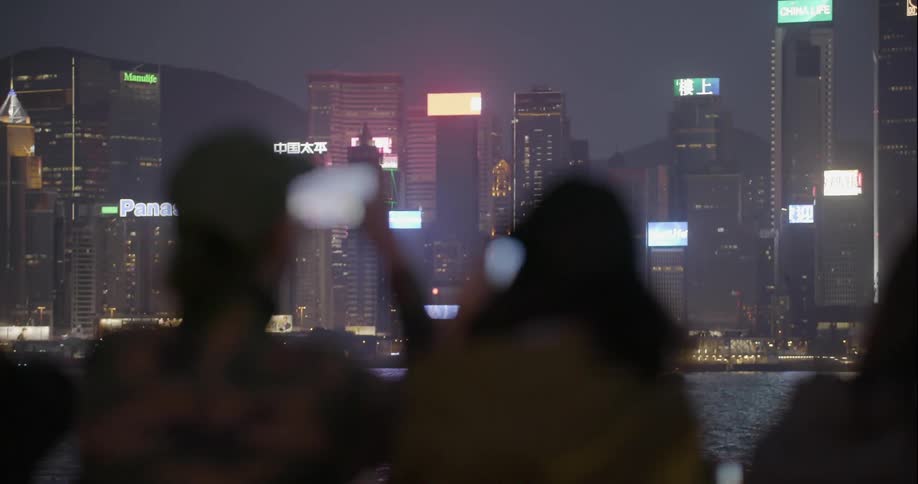 People standing at a railing and taking pictures of the large city across the river at night